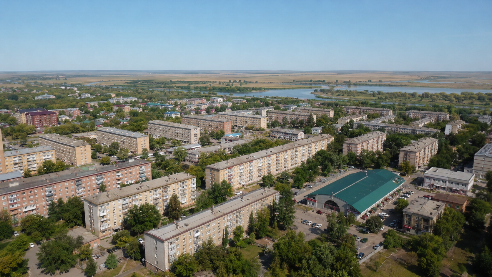 Вид с высоты на жилой район Петропавловска: пятиэтажки, дворы с деревьями и парковки, равнина Ишима и степь на горизонте под ясным небом